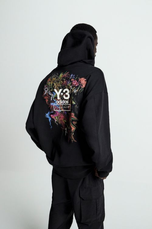 Back view of a person in a black Y-3 Adidas hoodie with a colorful graphic on the back, and black cargo pants in a neutral setting.