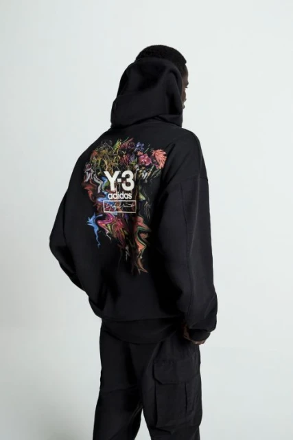 Back view of a person in a black Y-3 Adidas hoodie with a colorful graphic on the back, and black cargo pants in a neutral setting.