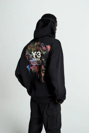 Back view of a person in a black Y-3 Adidas hoodie with a colorful graphic on the back, and black cargo pants in a neutral setting.