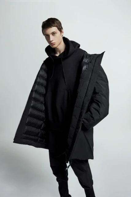 Studio shot of a male fashion model posing in a black hoodie and puffer coat, on a white background. The model looks at the camera.