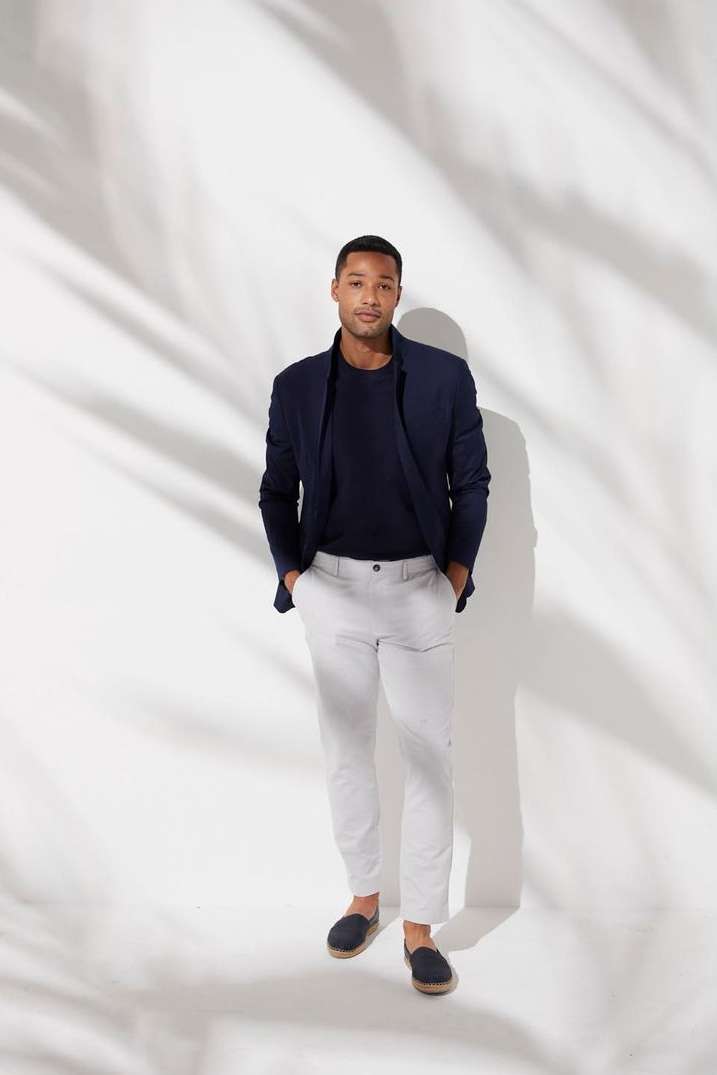 An adult male in a navy blazer, t-shirt and light trousers stands in a studio with soft lighting and shadows, hands in his pockets.