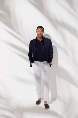 An adult male in a navy blazer, t-shirt and light trousers stands in a studio with soft lighting and shadows, hands in his pockets.