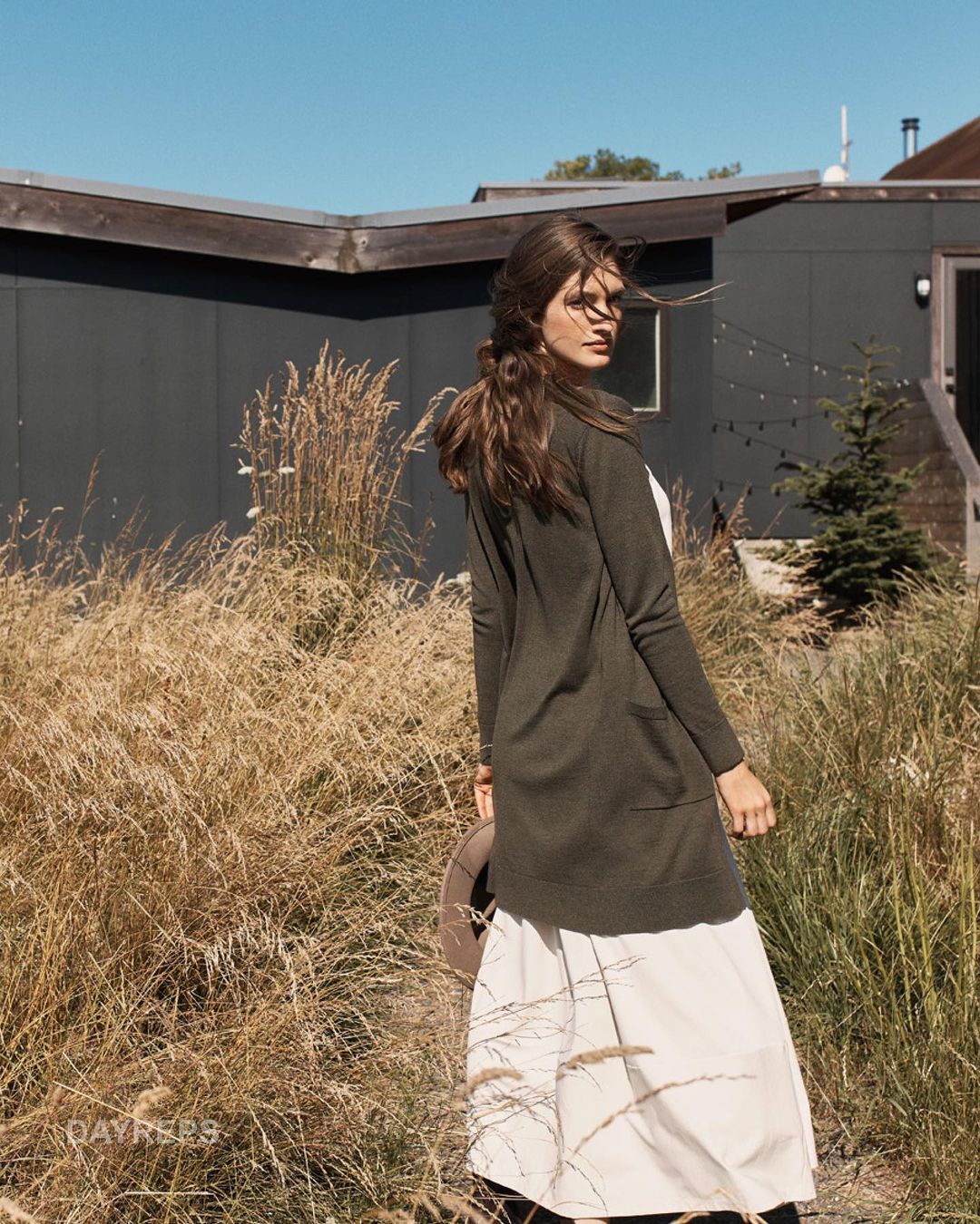 A woman in a cardigan and long dress stands in a field of tall grass near a modern grey house on a sunny day.