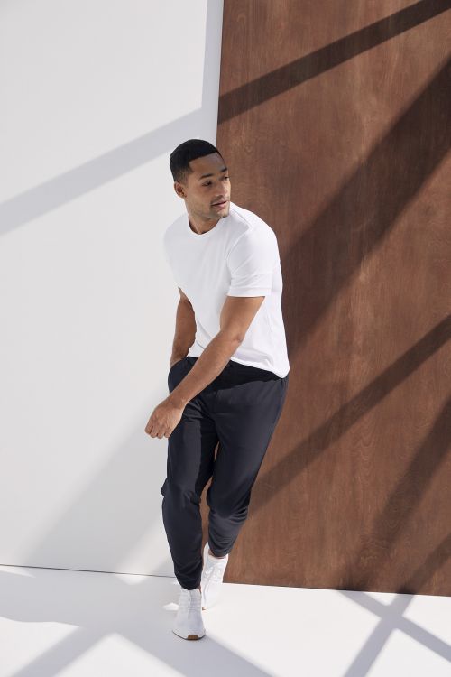 Athletic man in white t-shirt and black joggers poses in a studio with white and brown panels, lit by natural, diffused light.