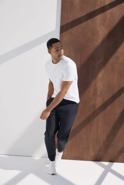 Athletic man in white t-shirt and black joggers poses in a studio with white and brown panels, lit by natural, diffused light.