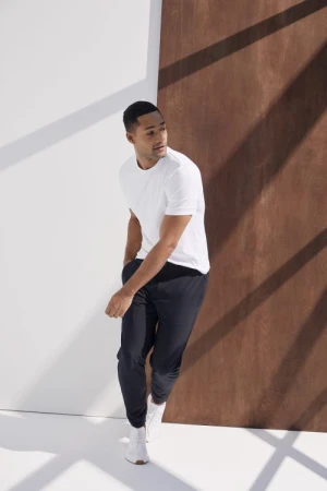 Athletic man in white t-shirt and black joggers poses in a studio with white and brown panels, lit by natural, diffused light.