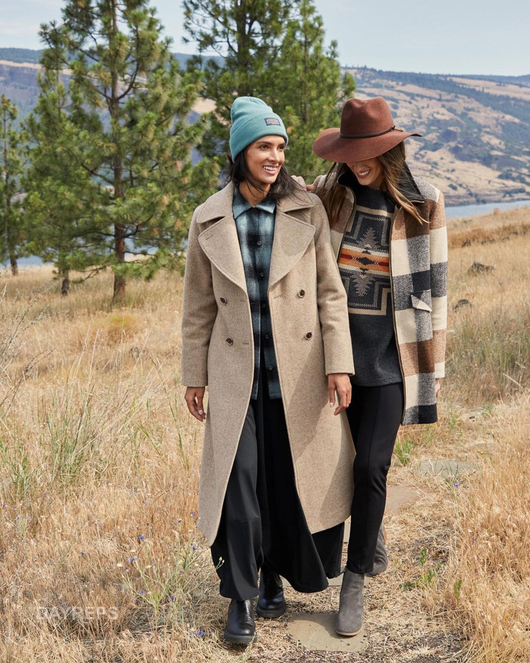 Two women stroll outdoors on a path, smiling and wearing hats, coats, and boots with a scenic, mountainous background.