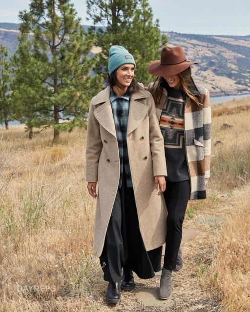 Two women stroll outdoors on a path, smiling and wearing hats, coats, and boots with a scenic, mountainous background.