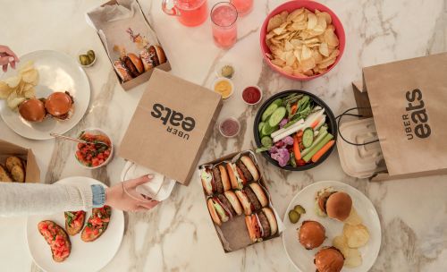 Overhead shot of a marble table laden with Uber Eats takeout containers, slider burgers, chips, veggie sticks, and pink drinks.
