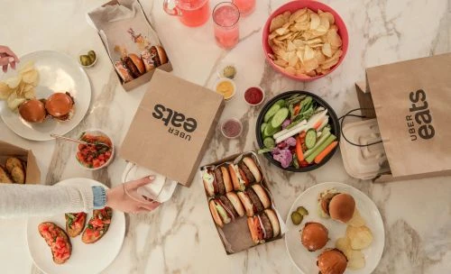 Overhead shot of a marble table laden with Uber Eats takeout containers, slider burgers, chips, veggie sticks, and pink drinks.