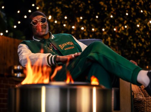 Snoop Dogg sitting outdoors in a green letterman jacket with Death Row Records text, fire pit, sunglasses, beanie, and fairy lights.