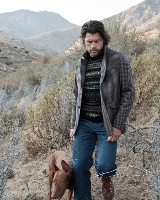 An adult male walking with a tan dog on a hillside trail, wearing a patterned sweater, jacket, and hiking boots, with mountains in the background.