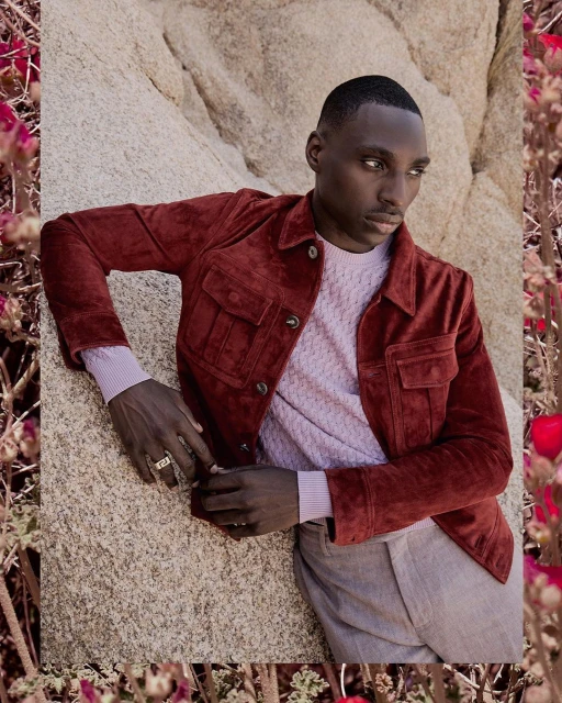 A black male model leans against a rock wearing a rust-colored suede jacket and light pink sweater, with floral framing.