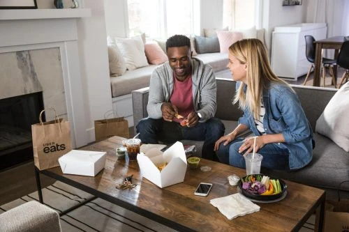 An African-American man and a Caucasian woman relax together on a sofa, enjoying a takeout meal from Uber Eats in a cozy living room.