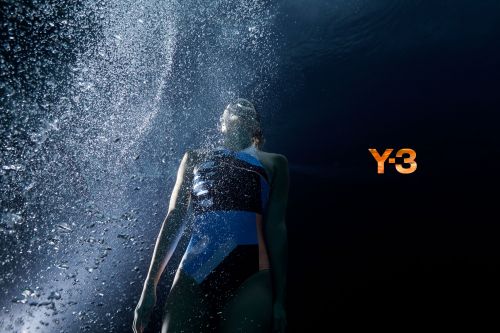 Underwater shot of a swimmer in a blue and black swimsuit surrounded by bubbles with the Y-3 logo visible against a black background.