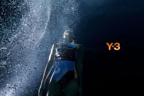 Underwater shot of a swimmer in a blue and black swimsuit surrounded by bubbles with the Y-3 logo visible against a black background.