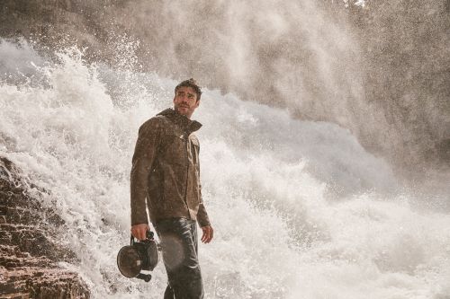 Adult male in jacket holding a vintage camera, standing in front of powerful waterfall, looking left. Outdoor setting, dramatic light.