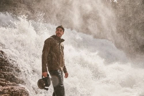 Adult male in jacket holding a vintage camera, standing in front of powerful waterfall, looking left. Outdoor setting, dramatic light.