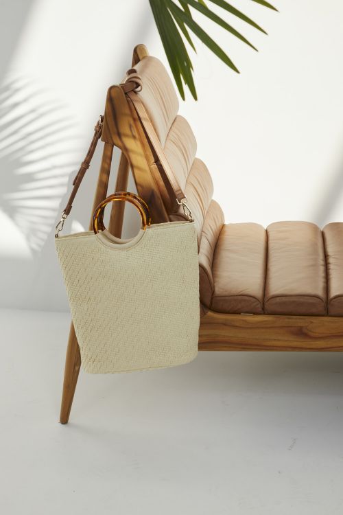 Stylish straw tote bag with bamboo handle casually draped over a light brown wood and cushioned chair with a white wall background.