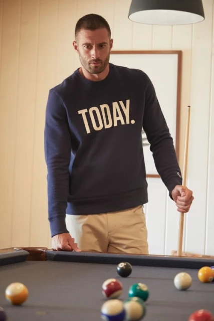 Indoor shot of a man in a sweatshirt holding a pool cue standing near a pool table with scattered billiard balls in a casual setting.