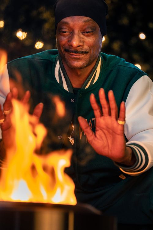 Snoop Dogg stands outdoors wearing a black beanie and green and white jacket, warming his hands over an open fire pit with fairy lights.