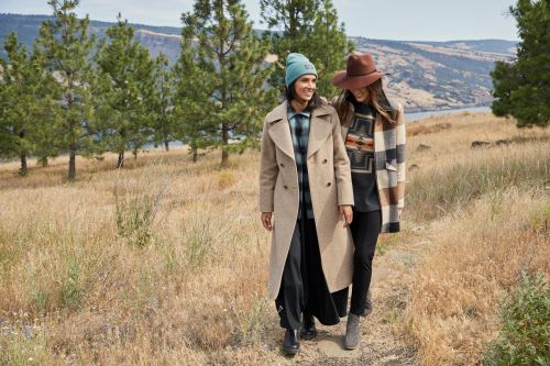 Two women in stylish coats and hats walk along a grassy path in a rural landscape with trees and distant hills under a bright sky.