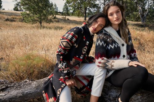 Two women in patterned sweaters and white and black leggings sit on a fallen log in a grassy field with trees in the background.