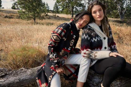Two women in patterned sweaters and white and black leggings sit on a fallen log in a grassy field with trees in the background.
