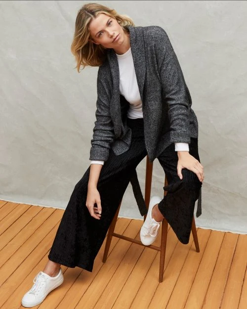 A fashionable adult woman with blonde hair, wearing a grey cardigan, white shirt, and black pants, sits on a wooden chair indoors.