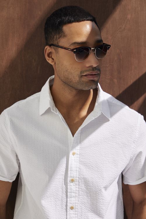 Close-up of a handsome adult man wearing sunglasses and a white shirt, posing indoors with shadow patterns on the background.