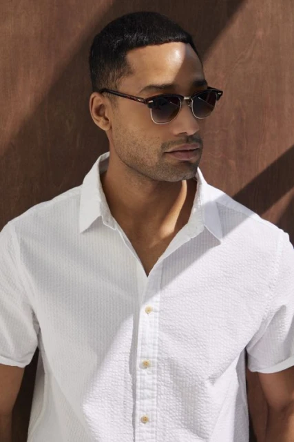 Close-up of a handsome adult man wearing sunglasses and a white shirt, posing indoors with shadow patterns on the background.