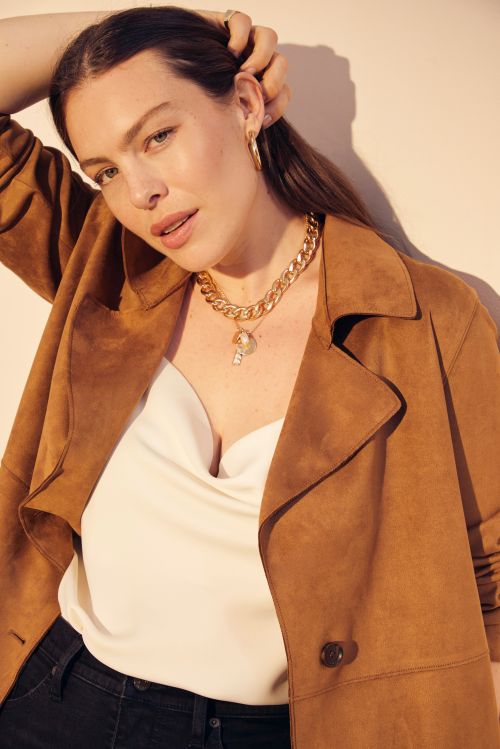 Portrait of a woman wearing a brown coat, white top, and gold necklace, looking at the camera in an indoor environment with light background.
