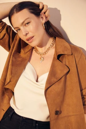 Portrait of a woman wearing a brown coat, white top, and gold necklace, looking at the camera in an indoor environment with light background.