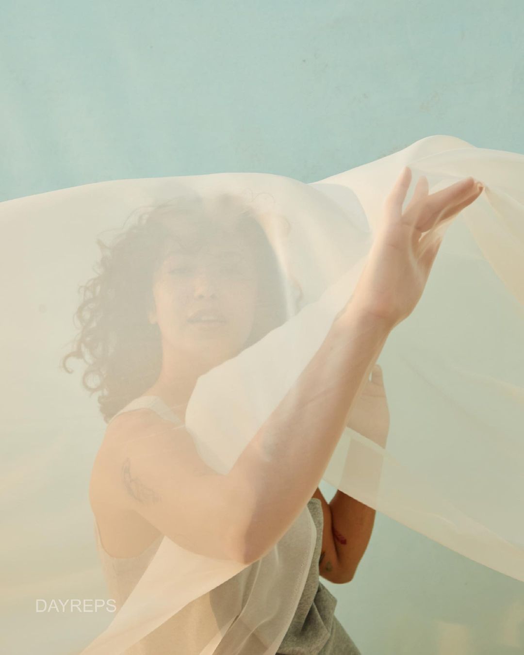 Woman with curly hair partially obscured by a white fabric, posing against a blue background in a portrait-style composition with studio lighting.