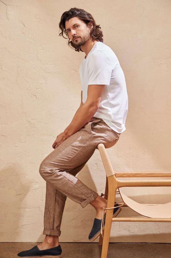 A man with long dark hair poses in a white t-shirt and brown pants. He sits on a light wood and woven chair in a minimalist style.