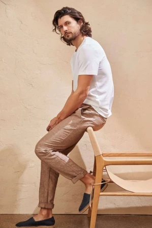 A man with long dark hair poses in a white t-shirt and brown pants. He sits on a light wood and woven chair in a minimalist style.