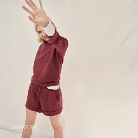 A woman smiles and poses in a maroon lounge set in front of a neutral backdrop, accessorized with a white undershirt and simple hair.
