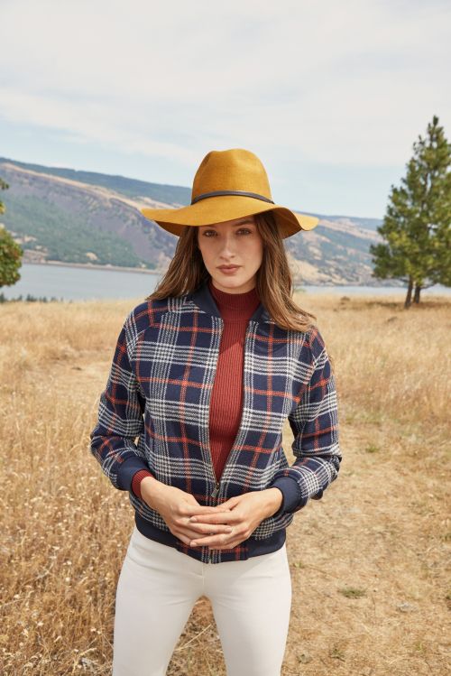 A fashionable woman posing outdoors, wearing a plaid jacket, white pants, and a wide-brimmed hat, with a scenic lake and hill backdrop.