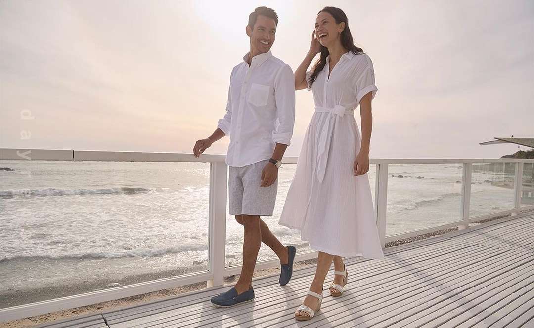 A couple strolls along a boardwalk by the ocean, both smiling and dressed in white, capturing a moment of seaside leisure.
