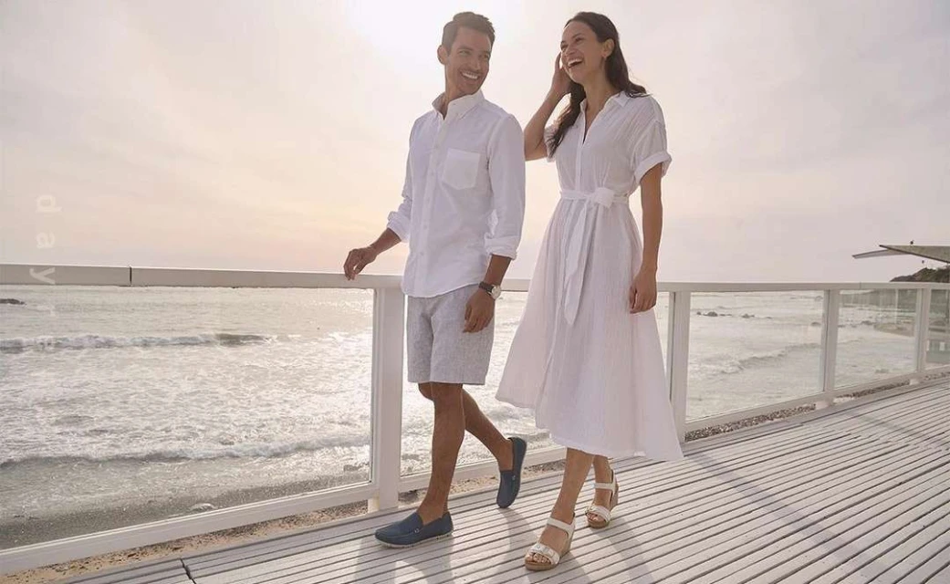 A couple strolls along a boardwalk by the ocean, both smiling and dressed in white, capturing a moment of seaside leisure.