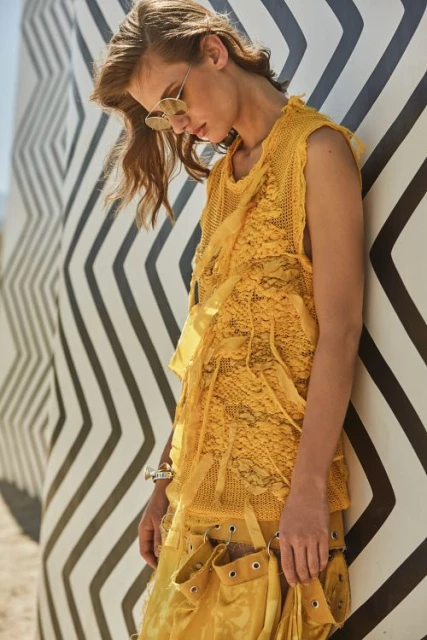 A stylish adult female poses in a yellow textured top and skirt leaning against a black-and-white patterned wall wearing sunglasses and bracelets.