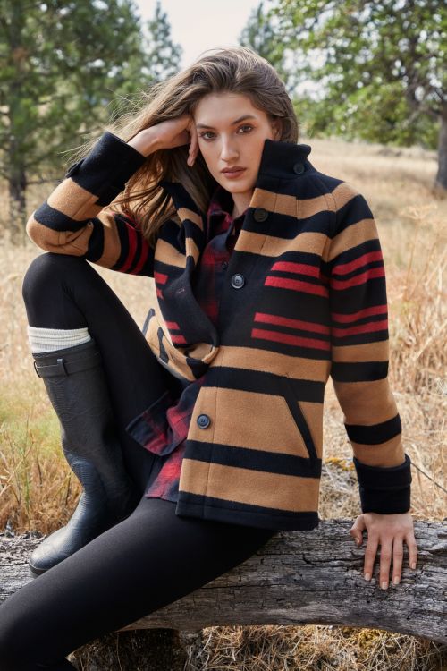 A woman in a striped coat sits on a log in a field of dry grass with trees in the background on a sunny day.