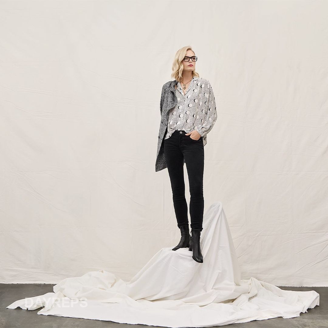 Blonde woman wearing glasses, patterned blouse, black jeans, and dark boots standing on a white draped fabric backdrop.