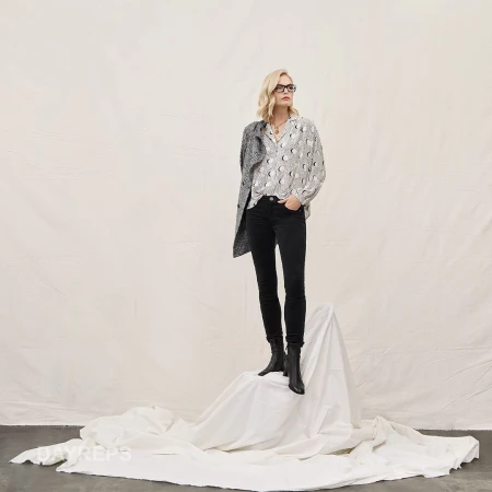 Blonde woman wearing glasses, patterned blouse, black jeans, and dark boots standing on a white draped fabric backdrop.