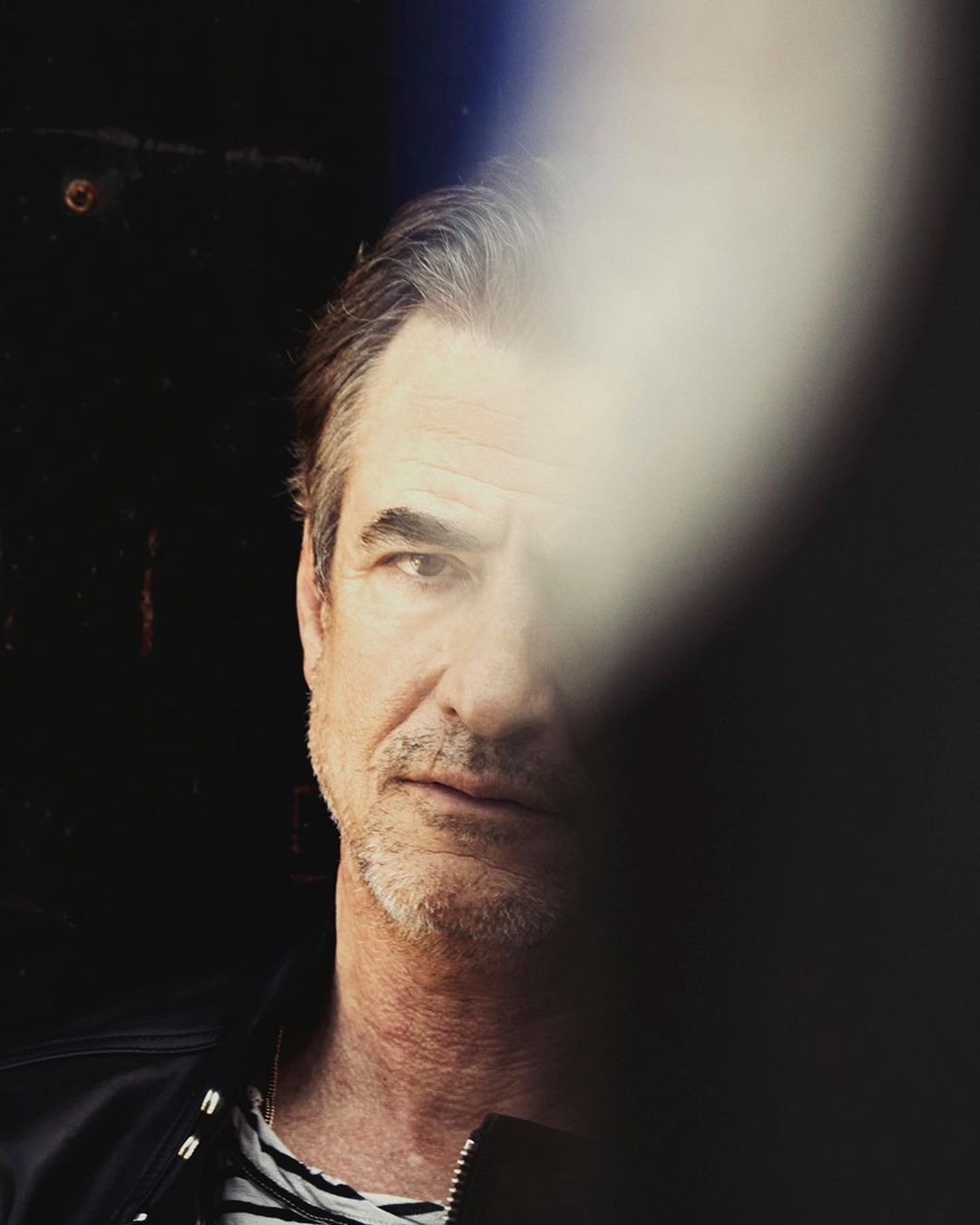 Portrait of a middle-aged adult man with graying hair in a dark setting, partially obscured by an out-of-focus white light.