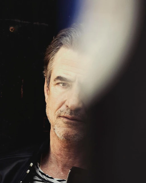 Portrait of a middle-aged adult man with graying hair in a dark setting, partially obscured by an out-of-focus white light.