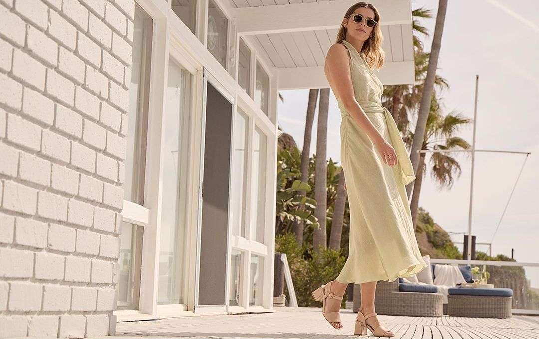 A woman in a light green dress and sunglasses standing on a porch with palm trees in the background on a sunny day.