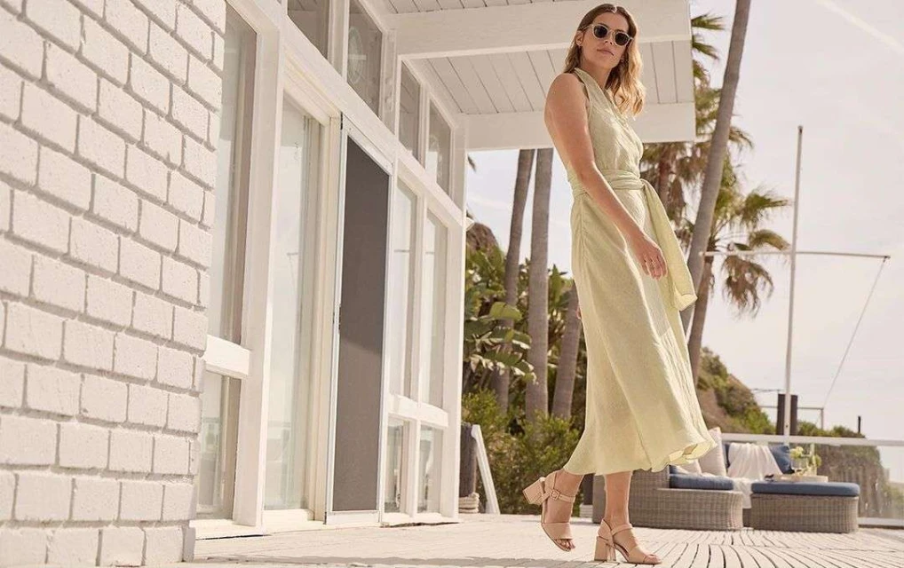 A woman in a light green dress and sunglasses standing on a porch with palm trees in the background on a sunny day.