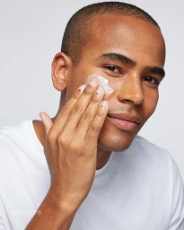 Close-up portrait of a Black man applying white skincare cream to his cheek, wearing a white t-shirt against a light grey background.