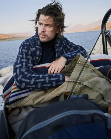 Man with dark hair wearing patterned blue cardigan and dark turtleneck leaning in the back of a vehicle near water